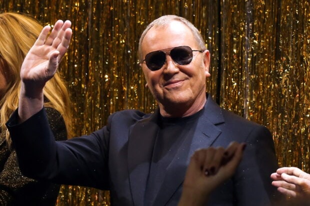 Michael Kors smiling wearing sunglasses and a dark suit in front of a gold backdrop