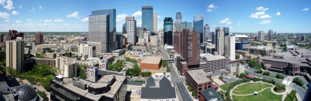 A panoramic view of the Minneapolis city skyline with tall skyscrapers and green spaces under a clear blue sky