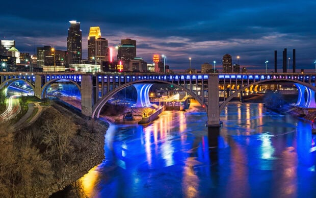 Nighttime view of Minneapolis skyline with a lit bridge over the river reflecting city lights