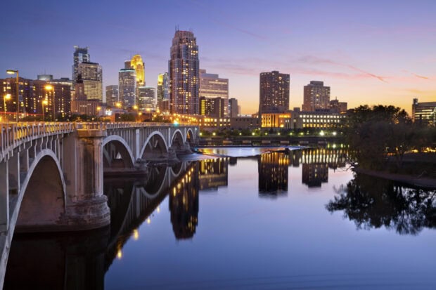 The Minneapolis city skyline with illuminated buildings and bridge reflected in the river at sunset