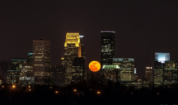 The Minneapolis cityscape with a large orange moon rising among skyscrapers at night