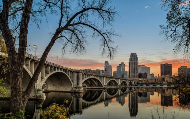 Historic Minneapolis bridge and city skyline reflected in calm river at sunset with trees in foreground