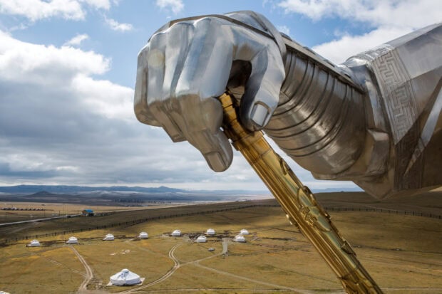 A large metallic hand sculpture holding a golden staff in the Mongolian steppe with traditional gers nearby
