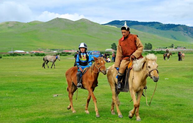 A young girl and a man riding horses on grassy plains in Mongolia with hills in the background