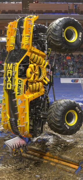 A yellow monster truck with spikes performing a stunt at an indoor arena with spectators