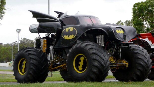A monster truck designed with Batman logos on its large tires and black body parked on grass