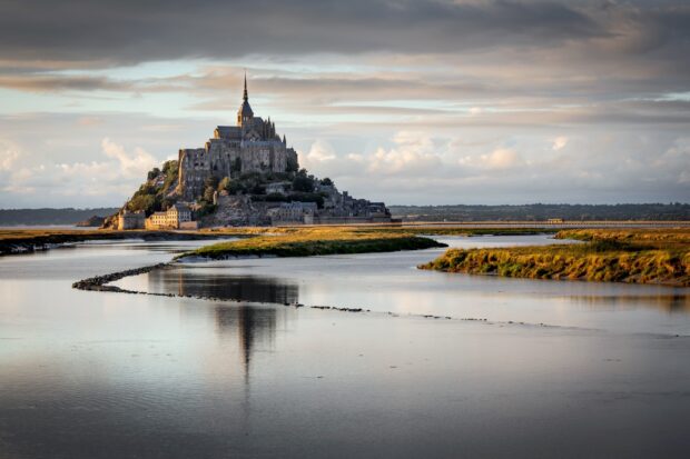 Mont Saint Michel viewed from a peaceful river landscape under a cloudy sky
