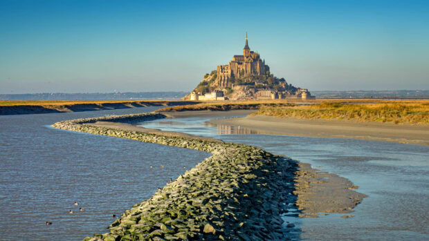 A scenic view of Mont Saint Michel with a winding stone path leading through the water and marshlands