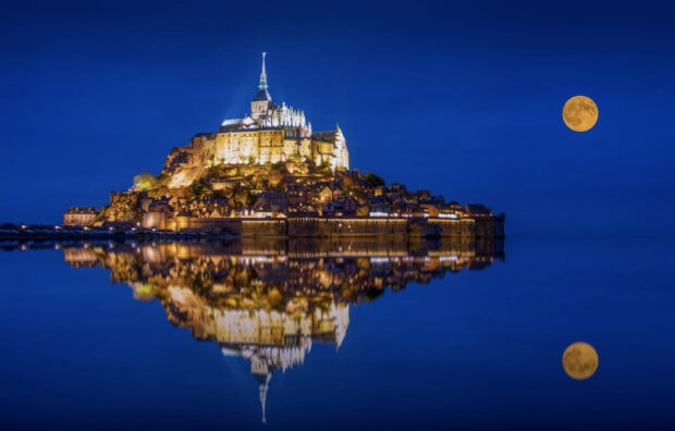 Mont Saint Michel castle illuminated at night with a full moon reflecting on calm water