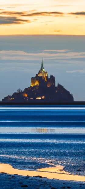 Mont Saint Michel castle illuminated at dusk with calm sea and colorful sky in the background