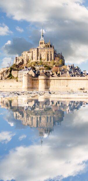 A stunning view of Mont Saint Michel reflected in calm water under a partly cloudy sky