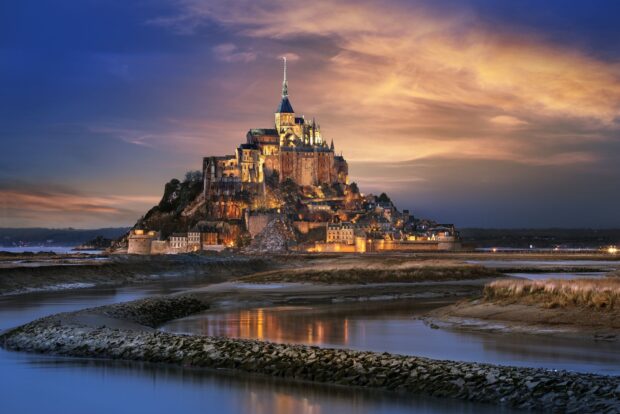 Mont Saint Michel at dusk with illuminated buildings and calm waters reflecting the sky
