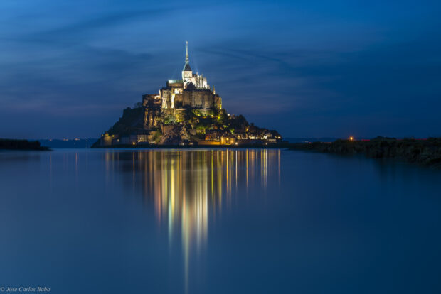 Mont Saint Michel reflected on calm water at night with illuminated buildings and clear sky