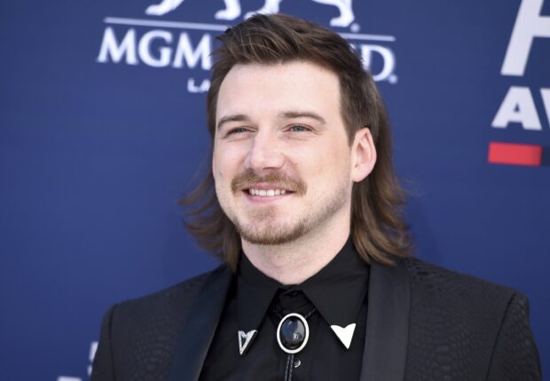 Morgan Wallen smiling with styled hair at a music event on a blue background