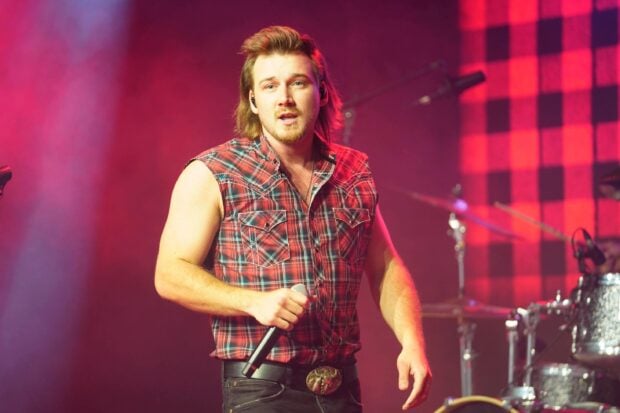 Morgan Wallen performing on stage wearing a sleeveless plaid shirt and holding a microphone during a live concert