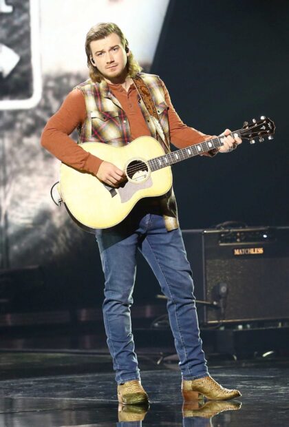 Morgan Wallen playing guitar on stage wearing a plaid vest and jeans in a live performance