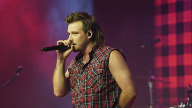 Morgan Wallen singing on stage with a plaid sleeveless shirt and microphone in hand