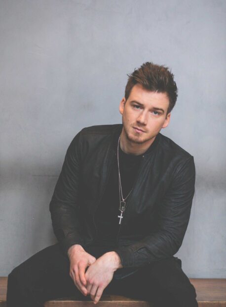 Young man wearing a leather jacket and necklace sitting against a gray wall Morgan Wallen