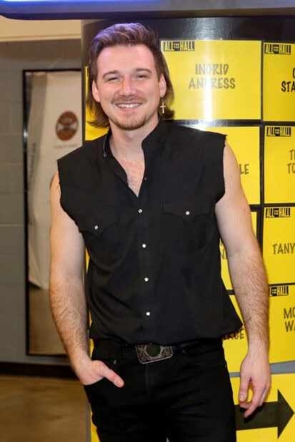 Morgan Wallen standing smiling in a black sleeveless shirt with an earring indoors