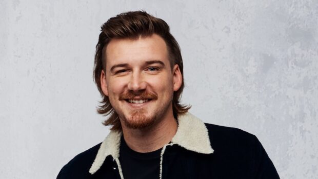 Morgan Wallen smiling with a mustache and beard in a casual jacket