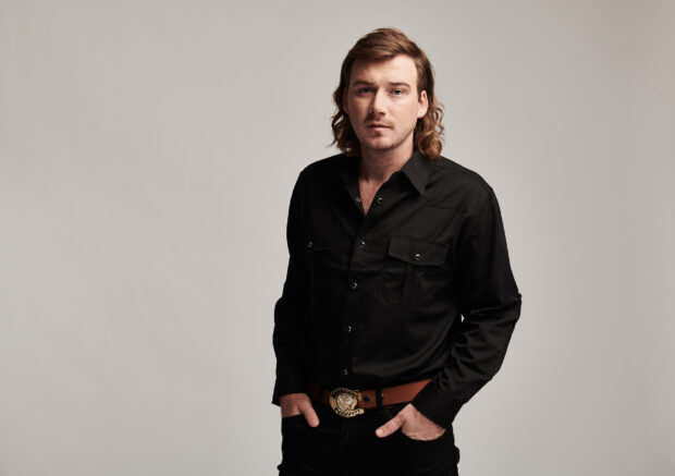 Morgan Wallen wearing a black shirt and brown belt standing against a plain background in studio lighting