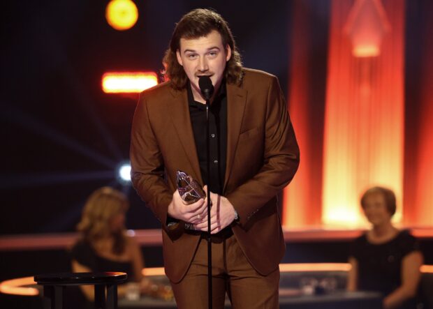 Morgan Wallen speaking at an award ceremony holding a trophy in a brown suit