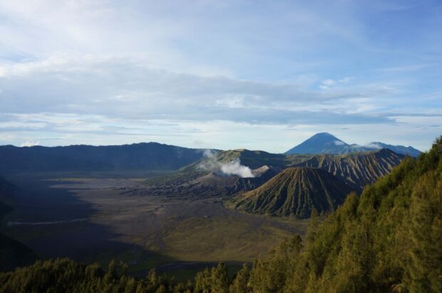 A scenic view of Mount Bromo volcano surrounded by lush green hills and a vast caldera under a blue sky