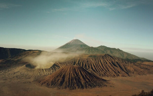 Mount Bromo volcano with smoke rising surrounded by rugged terrain and mountains under clear sky
