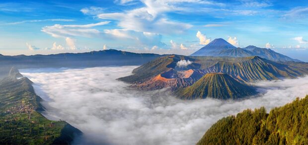 Volcanic landscape of Mount Bromo surrounded by clouds and hills under a bright blue sky