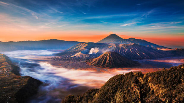 Volcanic landscape of Mount Bromo with smoke rising and surrounding mountains at sunrise
