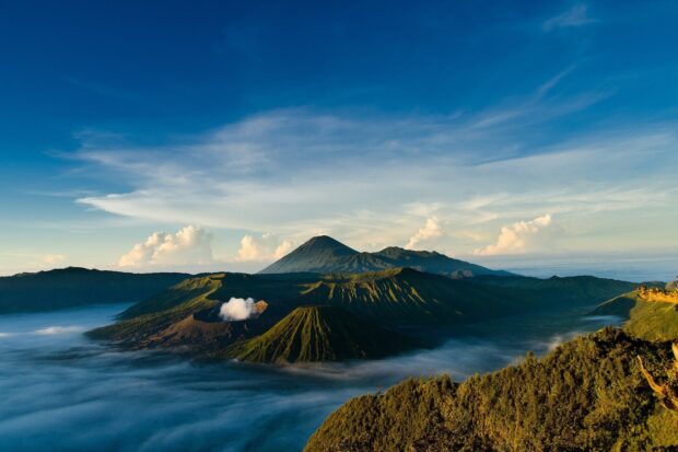 A scenic view of Mount Bromo volcano surrounded by mist and green hills under a clear blue sky