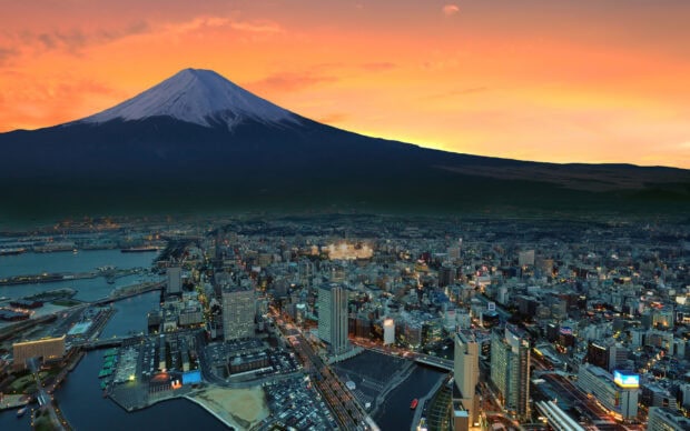 A cityscape at dusk with Mount Fuji looming in the background under an orange sky