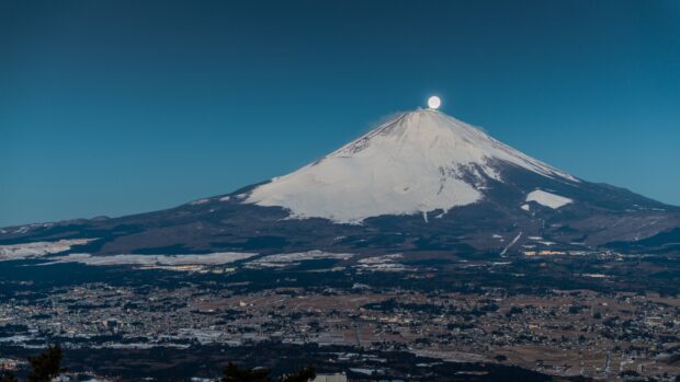 Snow covered Mount Fuji under a clear sky with the moon appearing above the peak