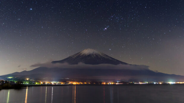 Mount Fuji stands tall under a starry night sky with reflections in the calm water below