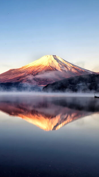Mount Fuji is reflected in a calm lake with mist surrounding the mountain at sunrise