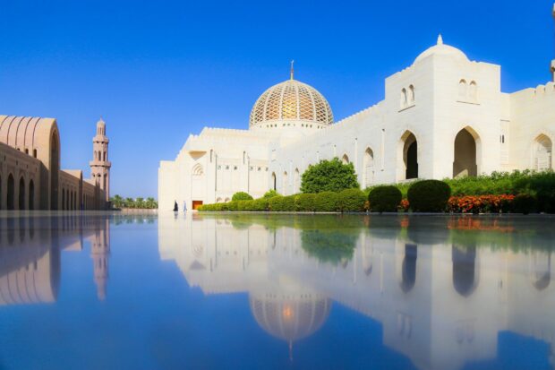 The Muscat architecture is reflected in the water under a clear blue sky