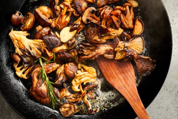 Various mushrooms cooking in a pan with a wooden spatula and fresh herbs