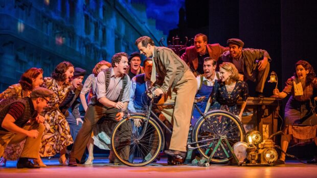 A lively musical scene with actors performing around a man on a vintage bicycle in a dramatic setting