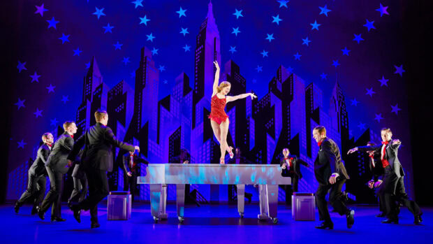 A musical performance featuring dancers and a singer on a grand piano on stage with city skyline backdrop