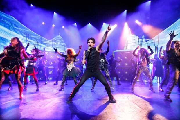 A musical performance with a lead singer and band on stage surrounded by colorful stage lights and dancers