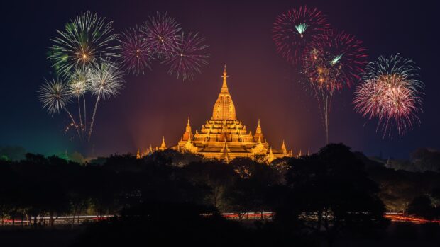 Golden Myanmar temple illuminated at night with fireworks in the sky above the landscape