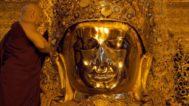 A monk placing gold leaf on a sacred statue in Myanmar temple interior