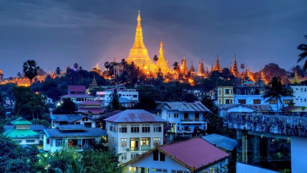 Golden pagodas in Myanmar cityscape at dusk with vibrant lights and tropical trees