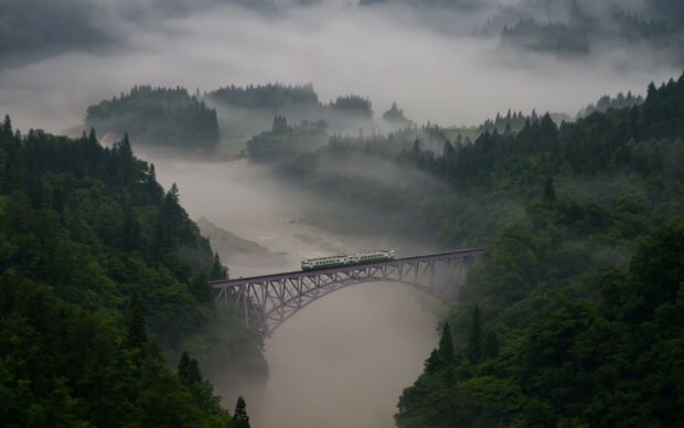 A scenic view of misty forest and river with a train crossing a bridge in nature National Geographic
