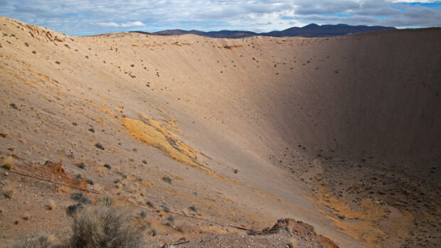A vast Nevada desert crater landscape with dry shrubs and distant mountains under a cloudy sky