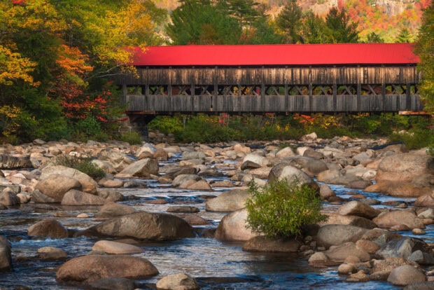 Covered bridge over rocky river in New Hampshire forest during autumn foliage