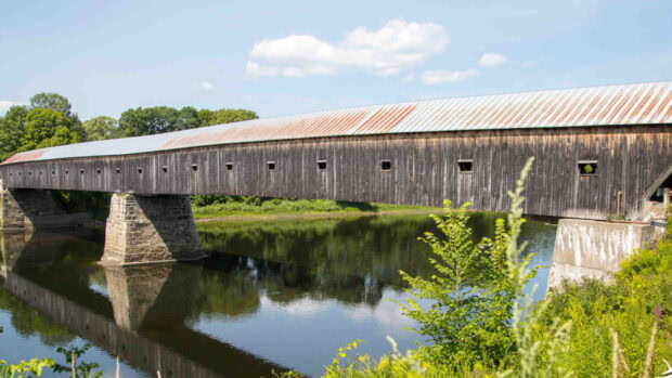 A historic wooden covered bridge over a calm river in New Hampshire surrounded by green trees