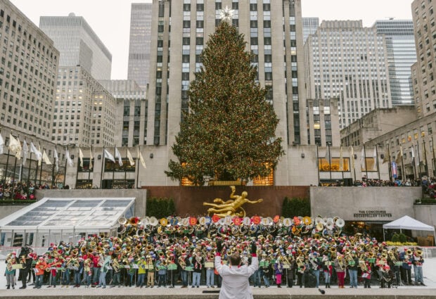 A large group of musicians performing in front of the New York Christmas tree at Rockefeller Center