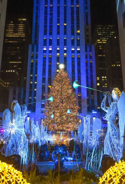 Large New York Christmas tree surrounded by angel sculptures and city buildings at night