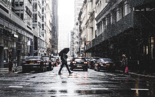 A person with an umbrella crossing a rainy New York street with cars parked along the buildings